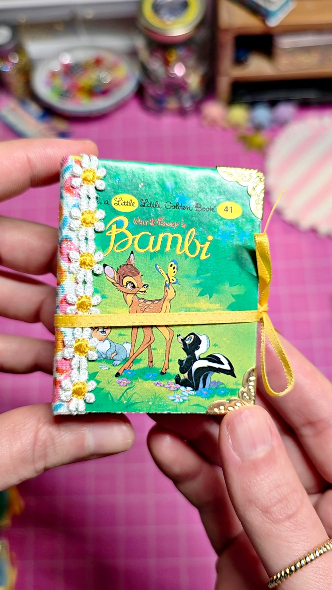A miniature paper book titled "Liz's Little Cotton Book: Bambi" held in two hands. The book cover features a green background with Bambi, a skunk, and daisies, and is tied with a yellow ribbon.
