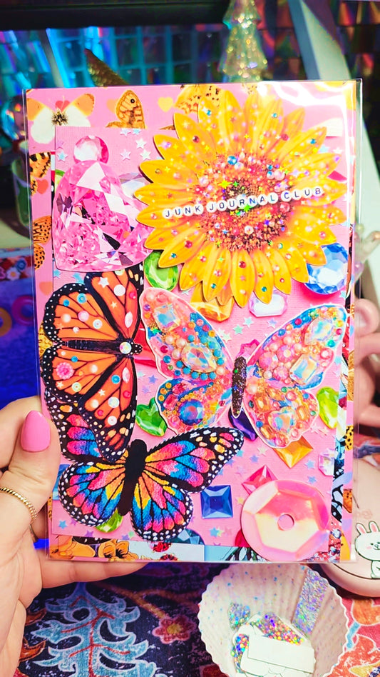 A hand holds a decorated pink notebook or journal cover featuring colorful butterflies, a large yellow flower, rhinestones, and decorative embellishments. The background is a colorful, slightly cluttered craft or desk setting.