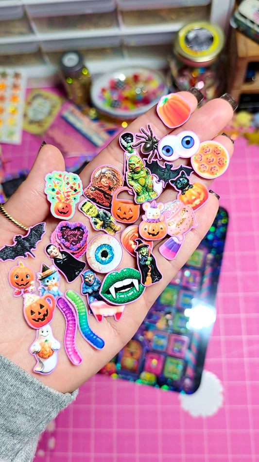 A hand holding a collection of colorful Halloween-themed stickers, including pumpkins, bats, eyeballs, a green monster, and a worm, over a pink cutting mat background.