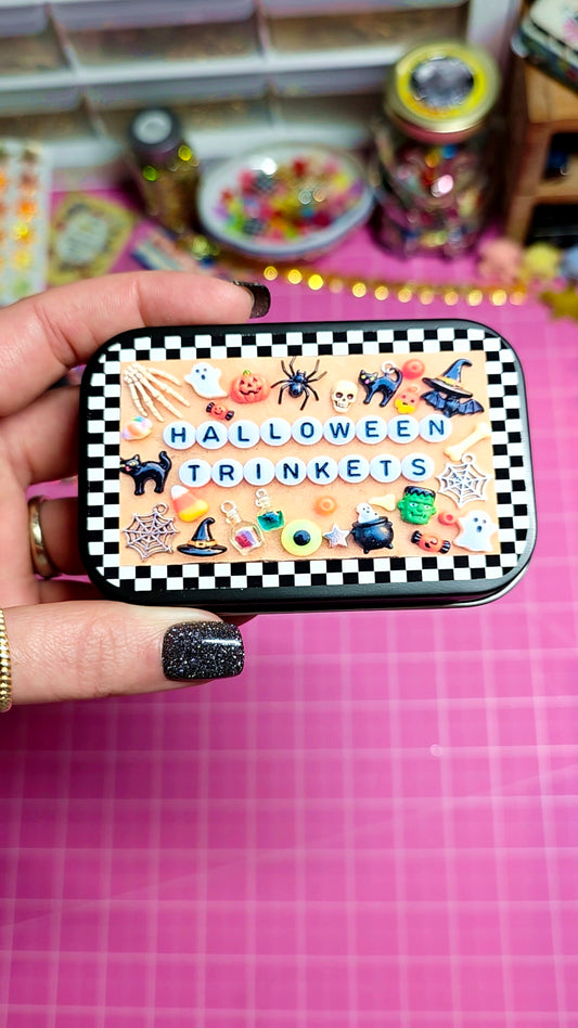 A hand with glittery black nail polish holds a rectangular tin of Halloween trinkets. The tin has a black and white checkered border and features various small Halloween illustrations like a spider, witch hat, potion bottle, and candy corn, with the text 'HALLOWEEN TRINKETS' in the center.