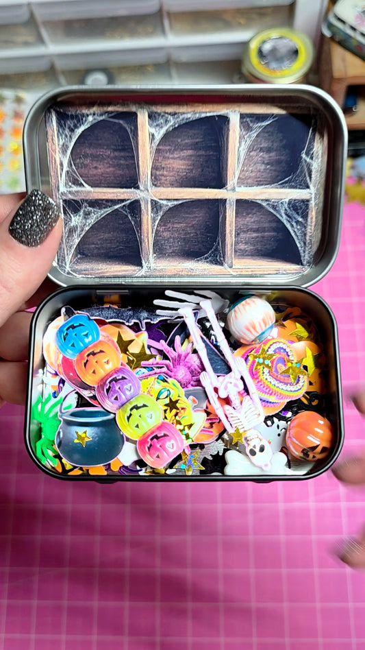 A hand holds an open metal tin filled with colorful Halloween-themed craft embellishments, including miniature pumpkins, ghosts, skeletons, and spiderweb decorations, against a pink grid background.