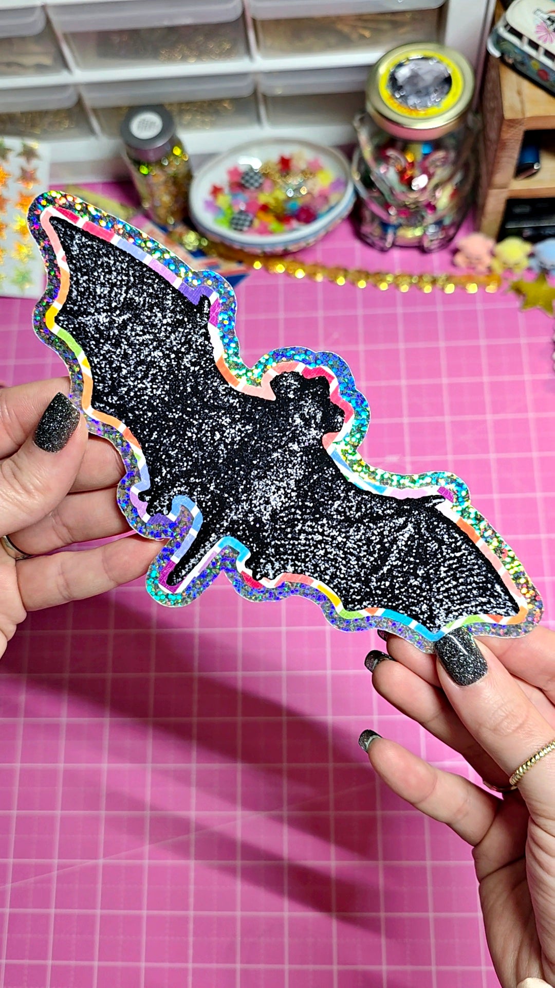 A person holds a black glitter bat-shaped sticker or patch with a holographic rainbow border over a pink cutting mat, with craft supplies visible in the background.