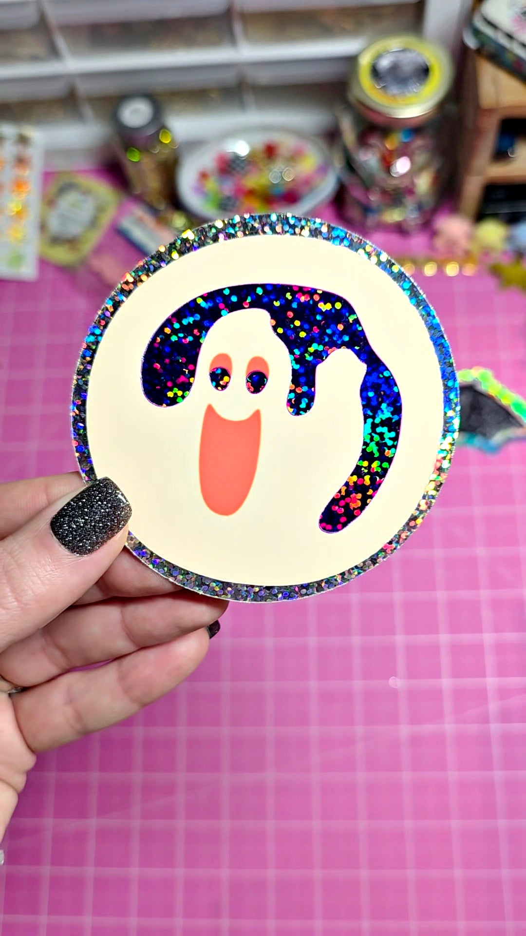 A hand with glittery black nail polish holds a round sticker featuring a ghost design. The sticker has a cream-colored center, a black and holographic glittery ghost body, and a glittery silver border.