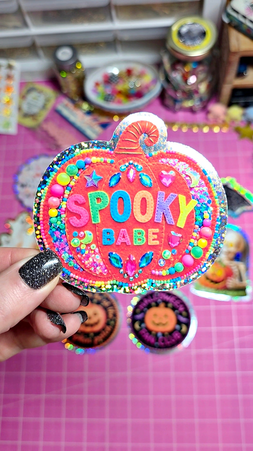 A hand with glittery black nail polish holds a round, pumpkin-shaped sticker or patch featuring the text 'SPOOKY BABE' in colorful letters on a pink glittery background. The scene is set on a pink cutting mat with other Halloween-themed items in the background.