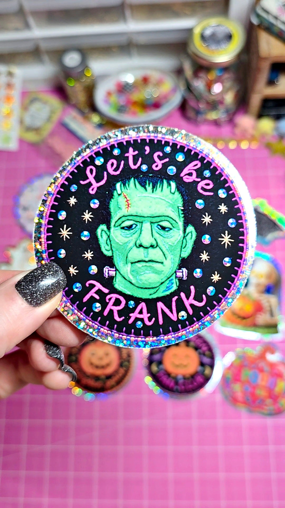 A hand with glittery black nail polish holds a round sticker featuring a green Frankenstein's monster face on a black background with the text 'Let's be FRANK' in pink and blue glitter. The background shows a pink craft mat with other stickers and craft supplies.