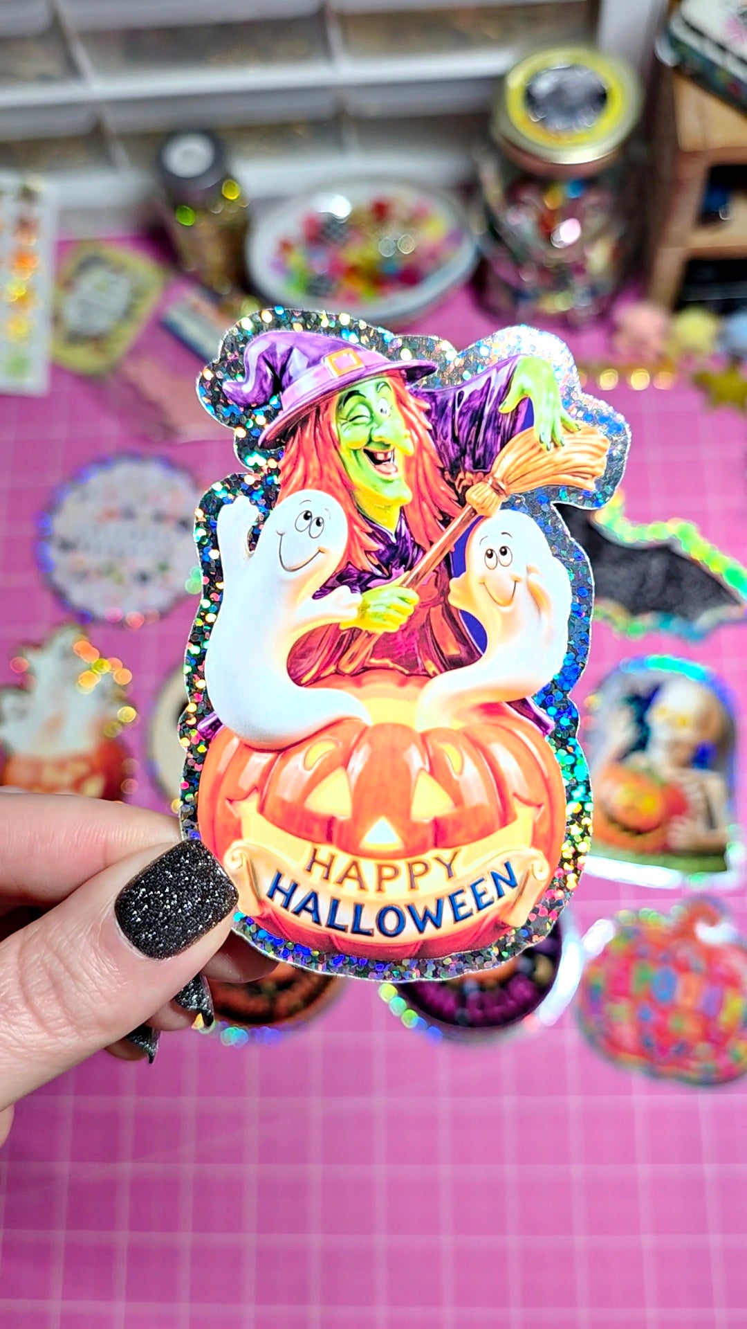 A hand with black glitter nail polish holds a holographic, iridescent Halloween sticker featuring a witch, ghosts, and a pumpkin with 'HAPPY HALLOWEEN' text, set against a pink grid background with other stickers.