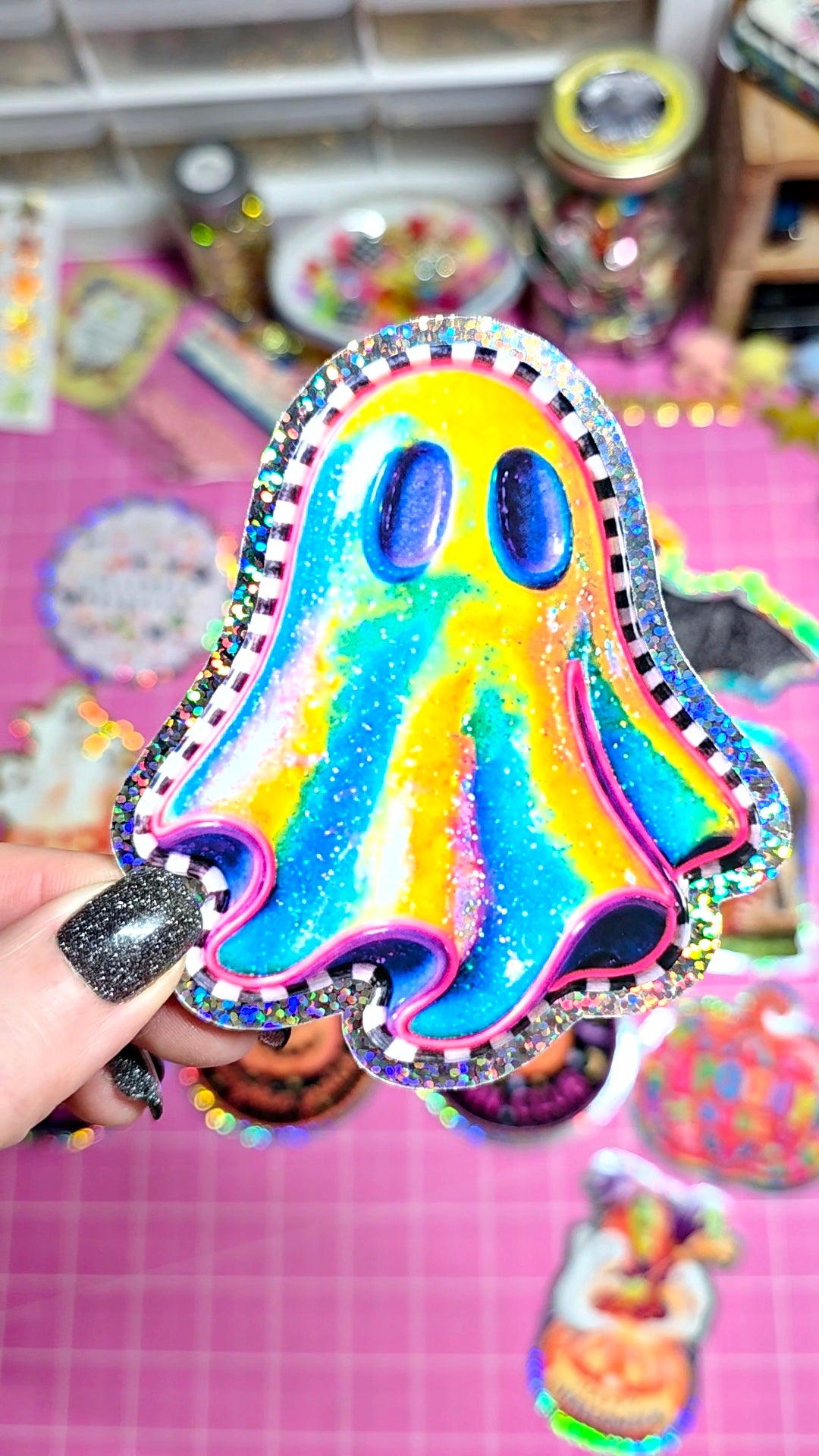 A hand with black glitter nail polish holds a colorful, holographic ghost-shaped sticker or patch with a glittery silver border. The background is a pink grid cutting mat with other craft supplies and stickers visible in a blurry, shallow depth of field.