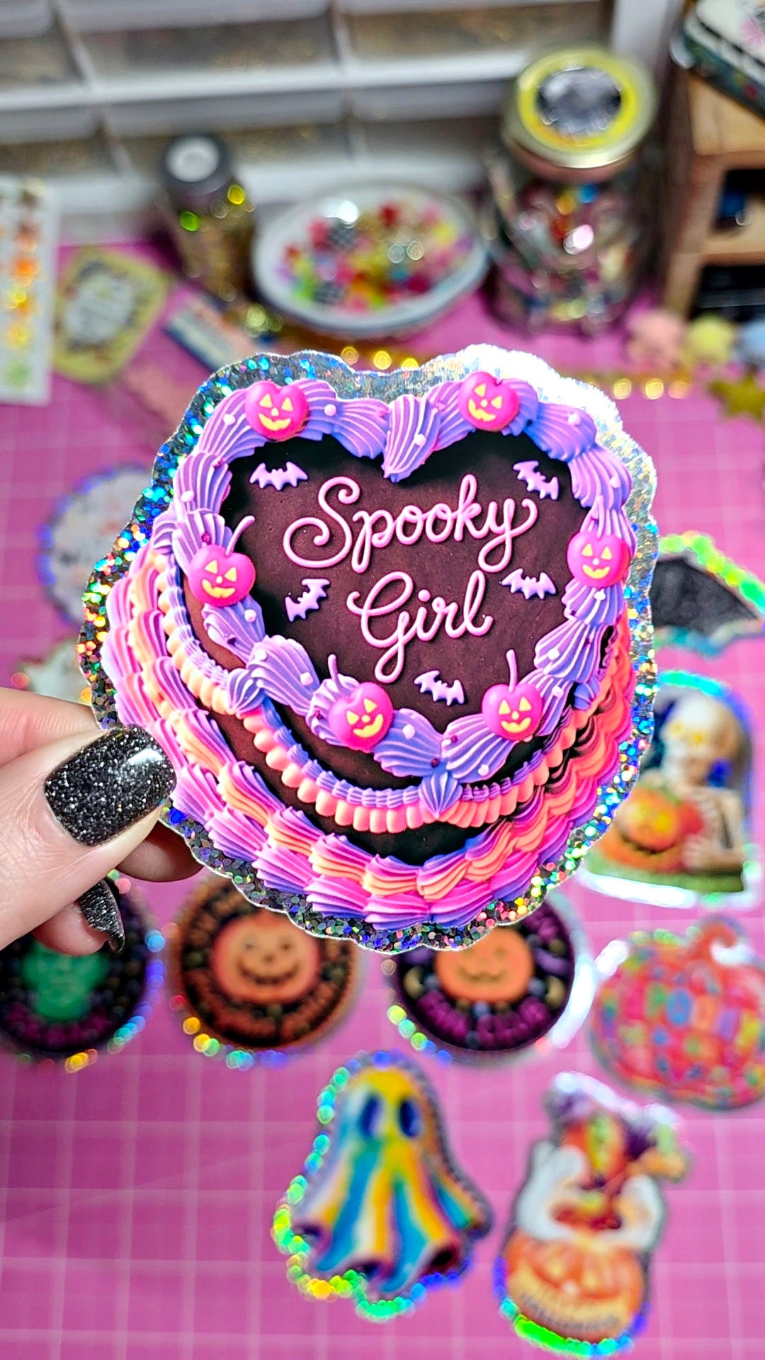 A hand with black glitter nail polish holds a die-cut sticker shaped like a frosted heart-shaped cake with the words 'Spooky Girl' in white script. The sticker features pink, purple, and dark brown frosting details, black bats, and small jack-o'-lantern faces.
