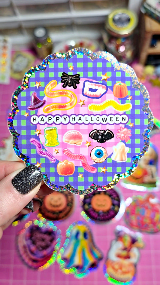 A hand with glittery black nail polish holds a scalloped holographic sticker featuring a purple and blue plaid background, candy illustrations, and the text 'HAPPY HALLOWEEN'. Other Halloween-themed stickers are scattered on a pink cutting mat in the background.