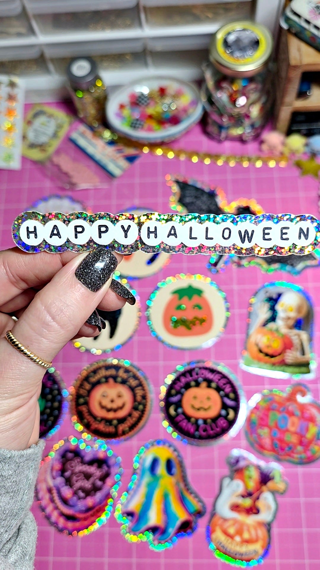 A hand with glittery black nail polish holds a holographic sticker that reads 'HAPPY HALLOWEEN' in white letters. The background is a pink grid surface covered with various colorful Halloween-themed stickers, including pumpkins, ghosts, and a witch.