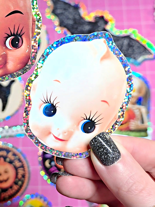 A hand with glittery black nail polish holds a die-cut sticker of a cartoon baby face with blue eyes and a holographic glitter border. Other stickers are visible in the background on a pink grid surface.