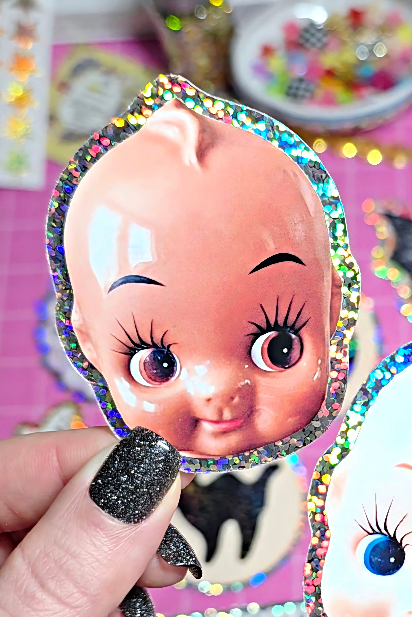A hand with glittery black nail polish holds a die-cut sticker or decal featuring a cute cartoon baby face with large eyes and a holographic glitter border. The background is a pink craft workspace with other stickers and supplies.