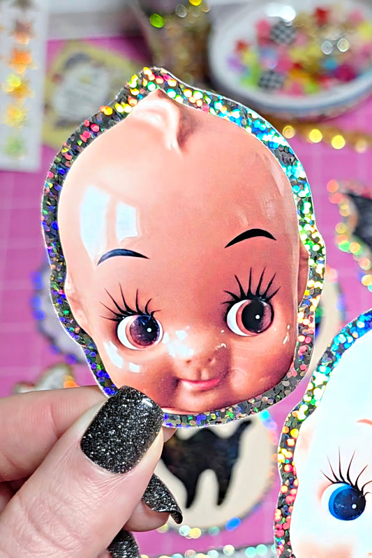 A hand with glittery black nail polish holds a die-cut sticker or decal featuring a cute cartoon baby face with large eyes and a holographic glitter border. The background is a pink craft workspace with other stickers and supplies.