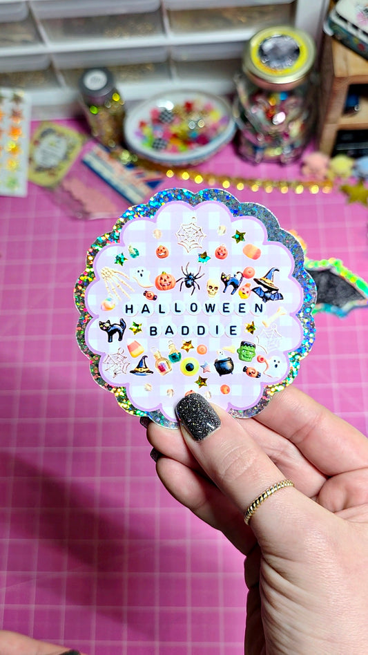 A hand with glittery black nail polish holds a scalloped white sticker sheet featuring Halloween-themed illustrations and the text 'HALLOWEEN BADDIE' against a pink cutting mat background.