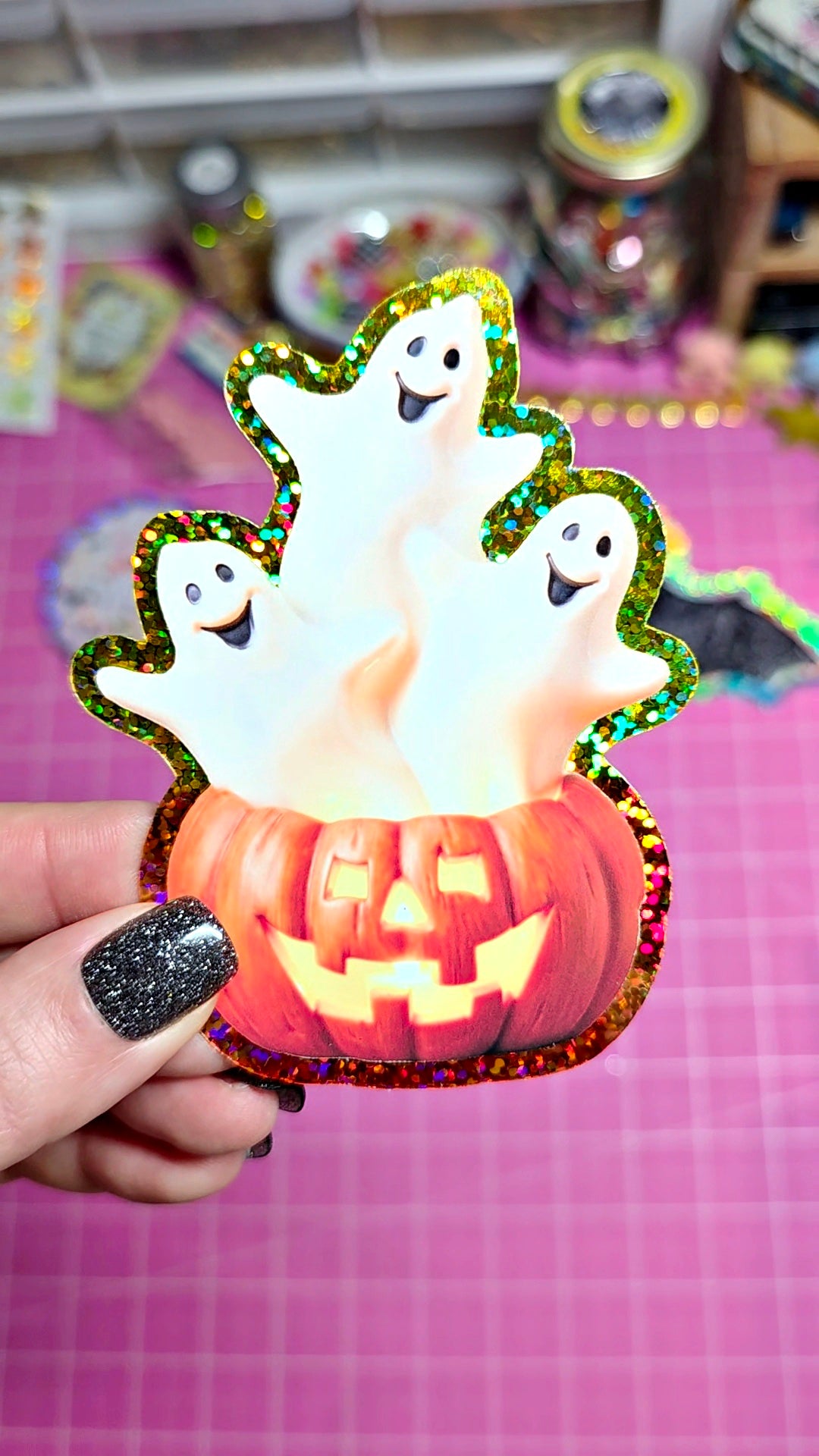 A hand with glittery black nail polish holds a die-cut Halloween sticker or decal featuring three white ghosts rising from a glowing orange jack-o'-lantern, with a holographic glitter border, against a pink grid background.