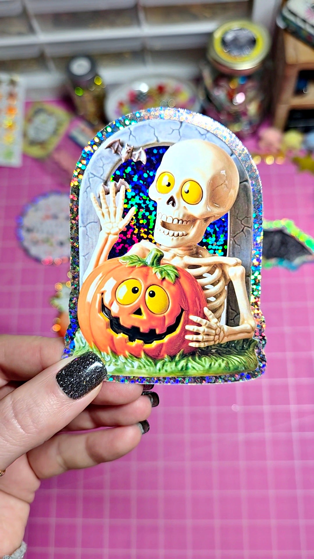 A hand with black glitter nail polish holds a holographic arch-shaped sticker featuring a cartoon skeleton and a jack-o'-lantern pumpkin against a pink cutting mat background.