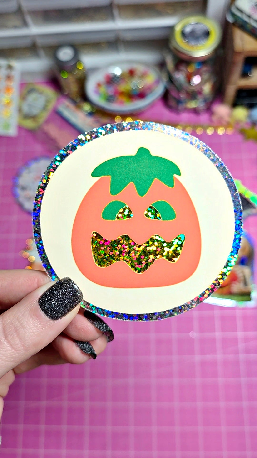 A hand with glittery black nail polish holds a round holographic sticker featuring a jack-o'-lantern face with a glittery gold bow tie, set against a pink grid background.