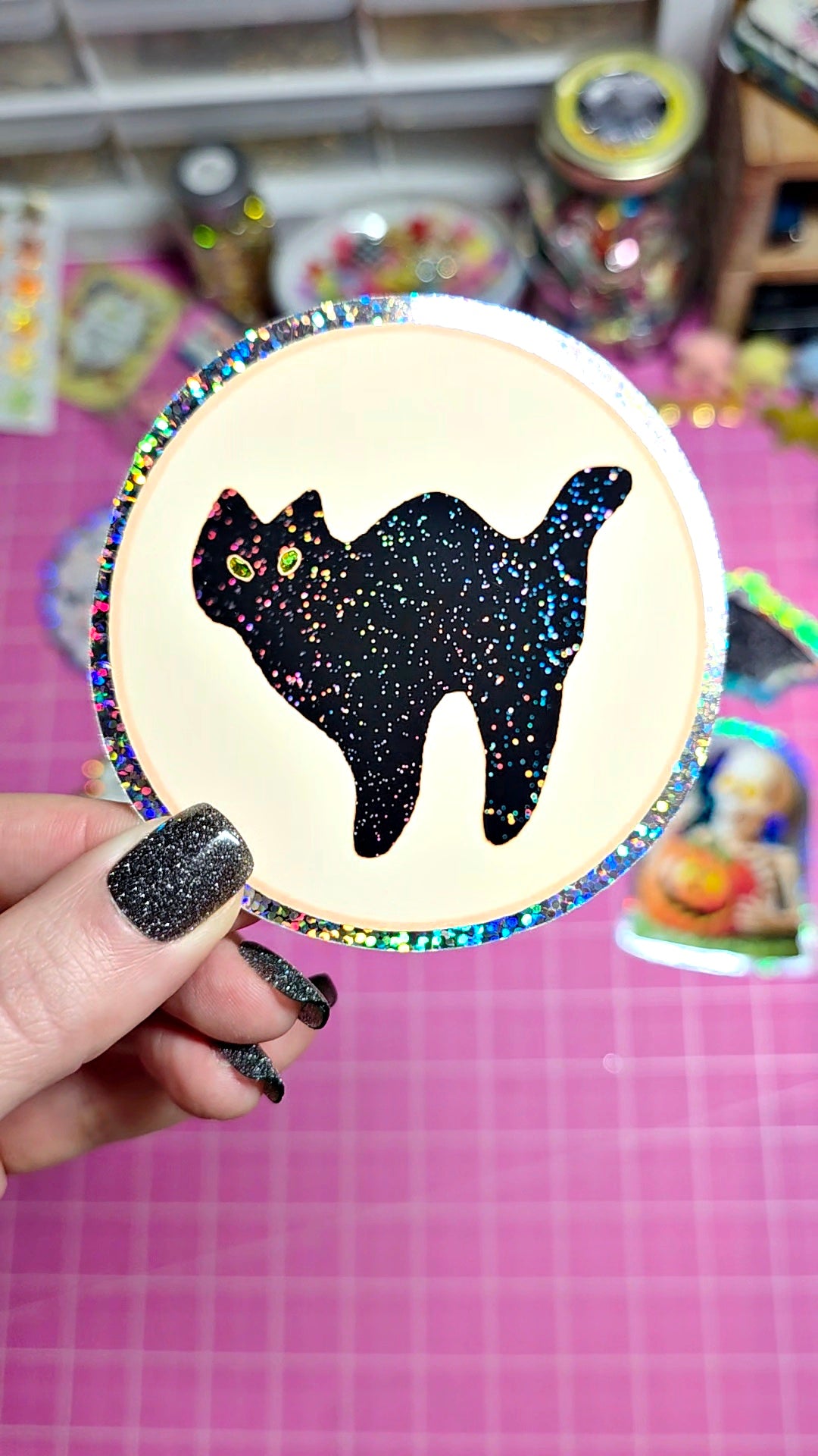 A hand with glittery black nail polish holds a round craft sticker or decal featuring a black cat silhouette filled with glitter, set against a pink grid cutting mat with craft supplies in the background.