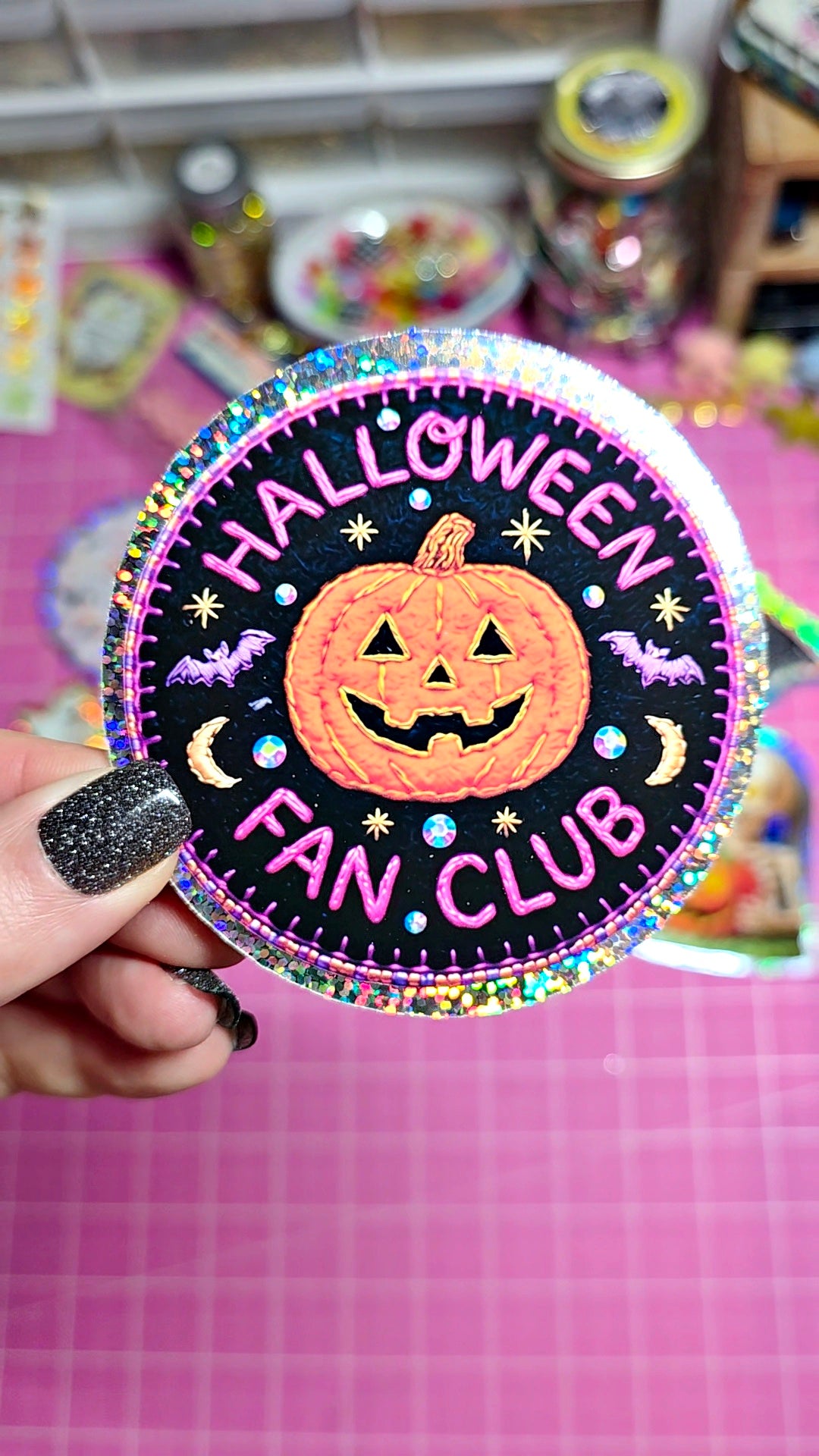 A hand with glittery black nail polish holds a round embroidered patch featuring a jack-o'-lantern and the text 'HALLOWEEN FAN CLUB' on a pink grid background.