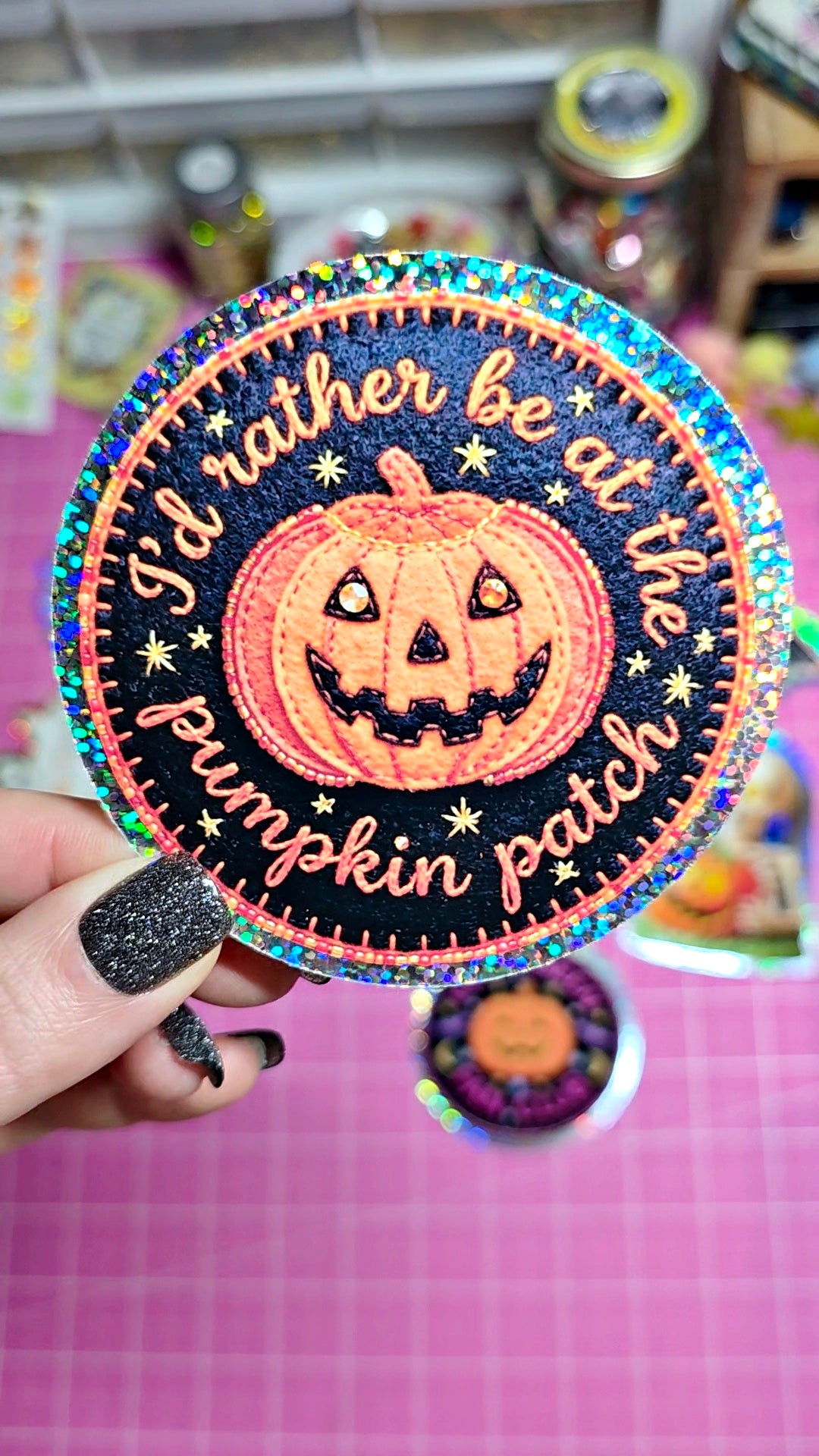 A hand with glittery black nail polish holds a round embroidered patch featuring a jack-o'-lantern and the phrase 'I'd rather be at the pumpkin patch' on a pink grid background.