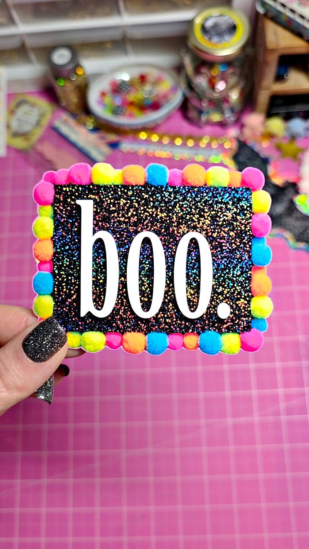 A hand with glittery black nail polish holds a square Halloween-themed card or sign featuring the word 'boo.' in white text on a black glitter background, framed by colorful pom-poms in pink, blue, yellow, green, and orange, set against a pink cutting mat and craft supplies.