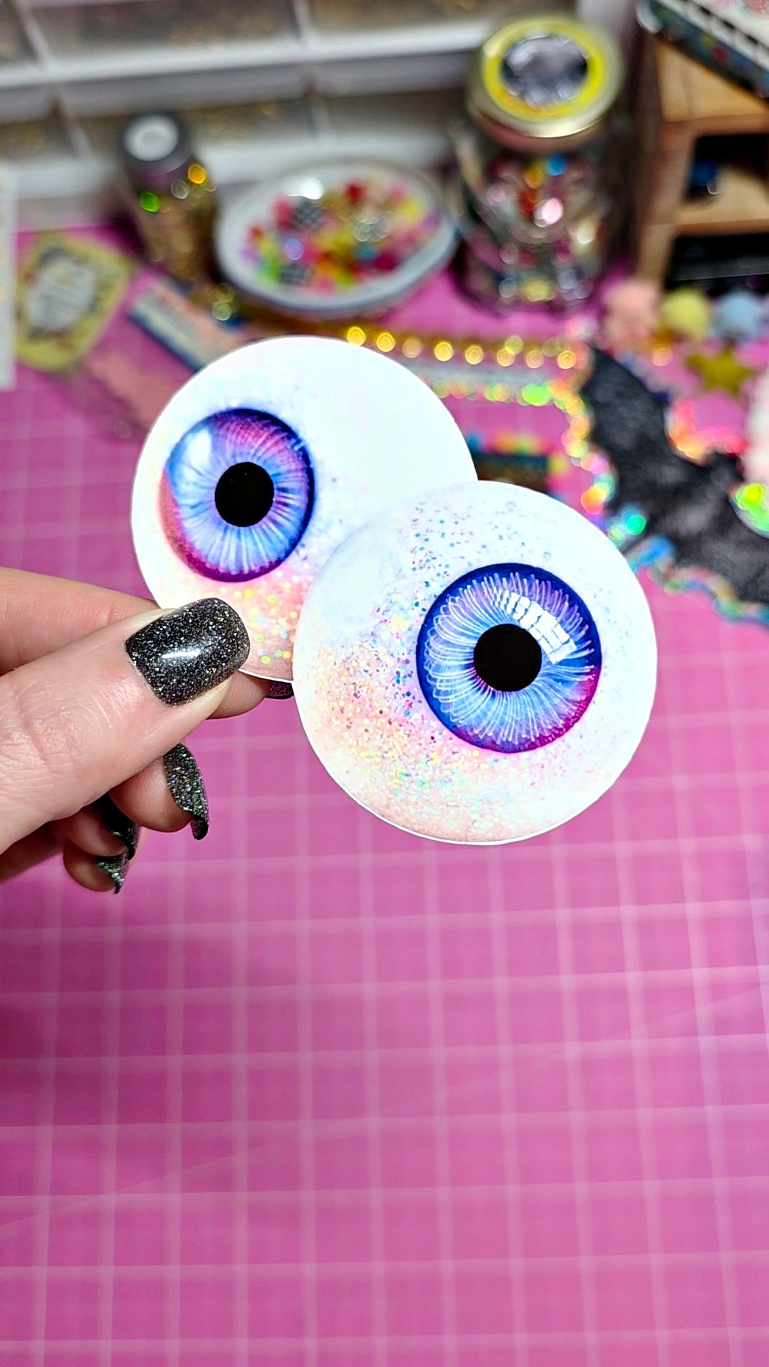 Two handmade, realistic plastic eyeball craft decorations held up by a hand with glittery black fingernails, set against a pink cutting mat background.