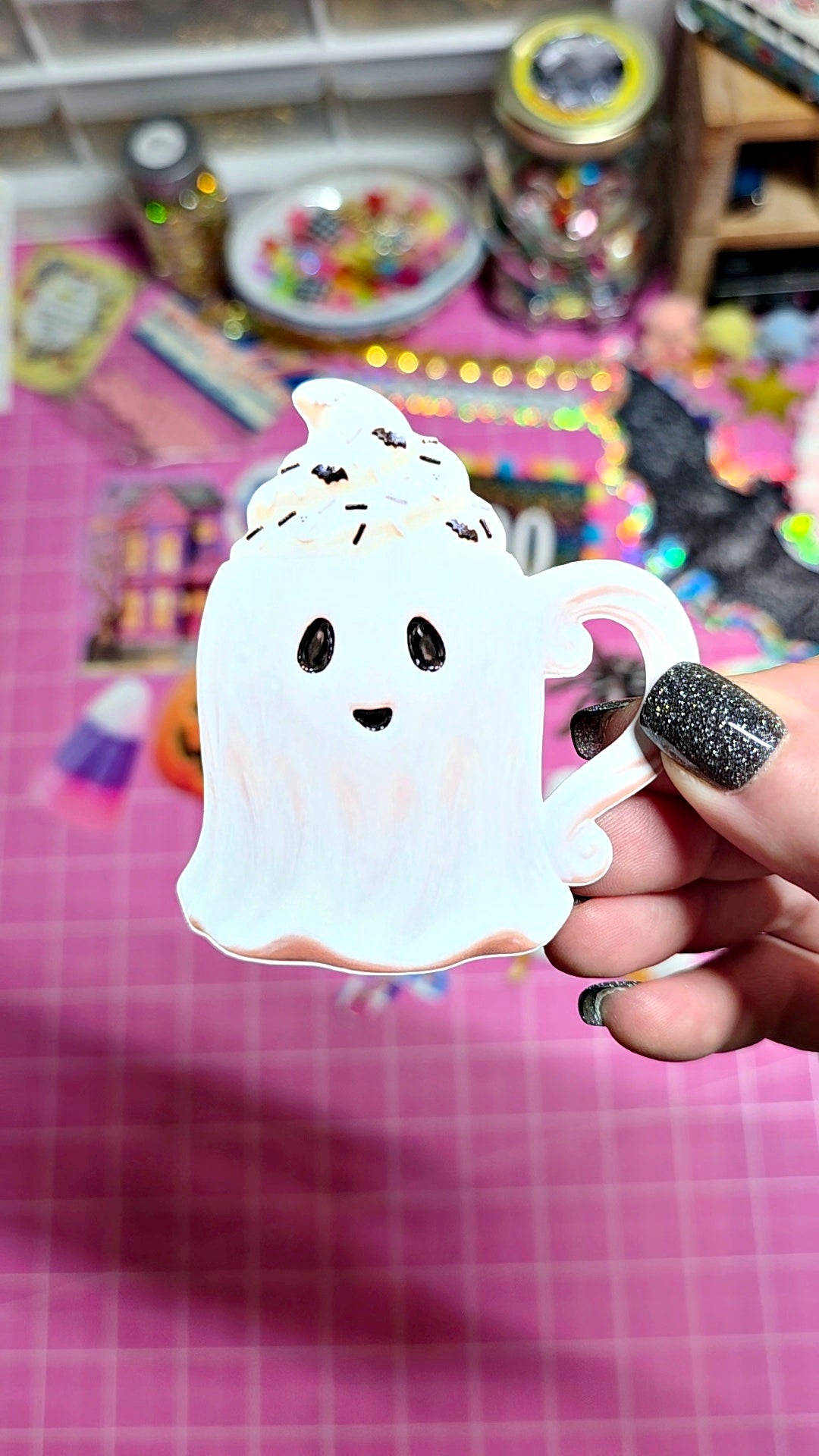A hand with glittery black nail polish holds a white ghost-shaped mug with a cute face, set against a pink craft mat and a blurred background of colorful supplies.