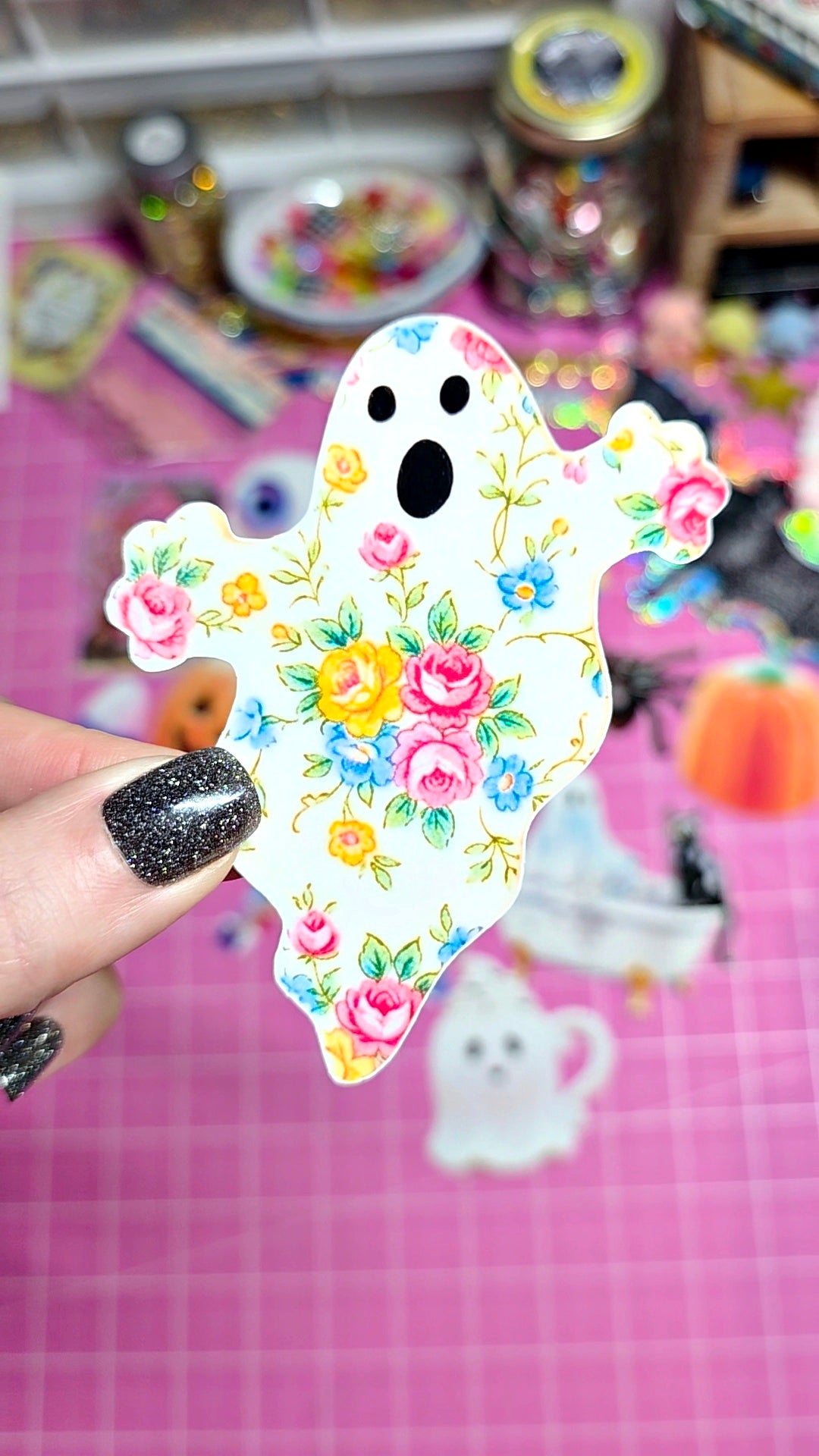 A hand with glittery black fingernails holds a die-cut ghost sticker featuring a colorful floral pattern on a white background. The scene is set against a pink cutting mat with other Halloween-themed stickers and craft supplies blurred in the background.