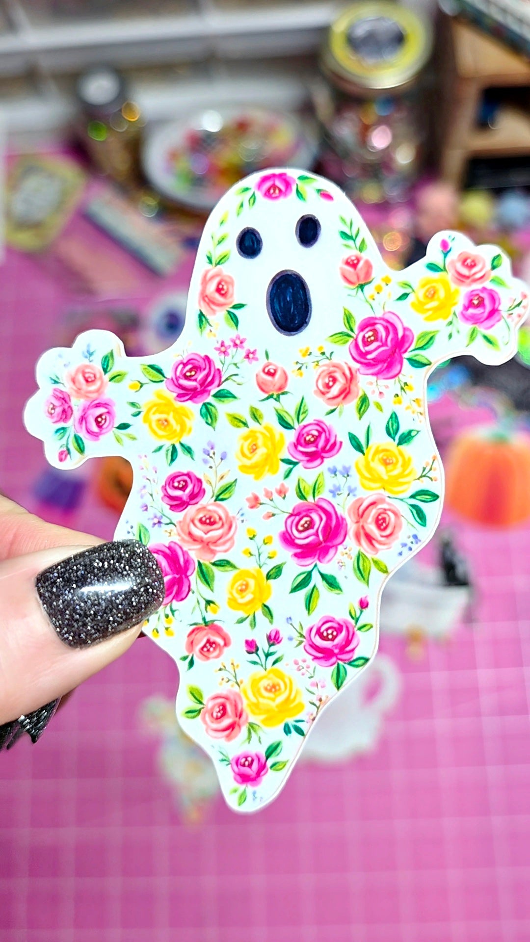 A hand with glittery black nail polish holds a die-cut ghost-shaped sticker or ornament decorated with a colorful floral pattern of pink, yellow, and white roses with green leaves, set against a pink grid background.