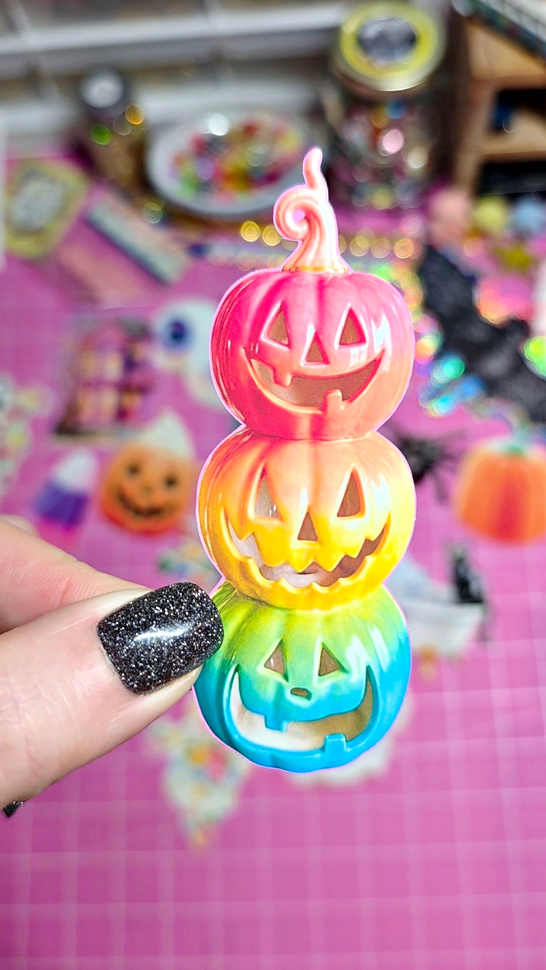A hand with glittery black nail polish holds a three-tiered stack of colorful jack-o'-lantern charms. The charms feature a pink top pumpkin, a yellow-middle pumpkin, and a blue-green bottom pumpkin, all with carved faces and a rainbow gradient transition.
