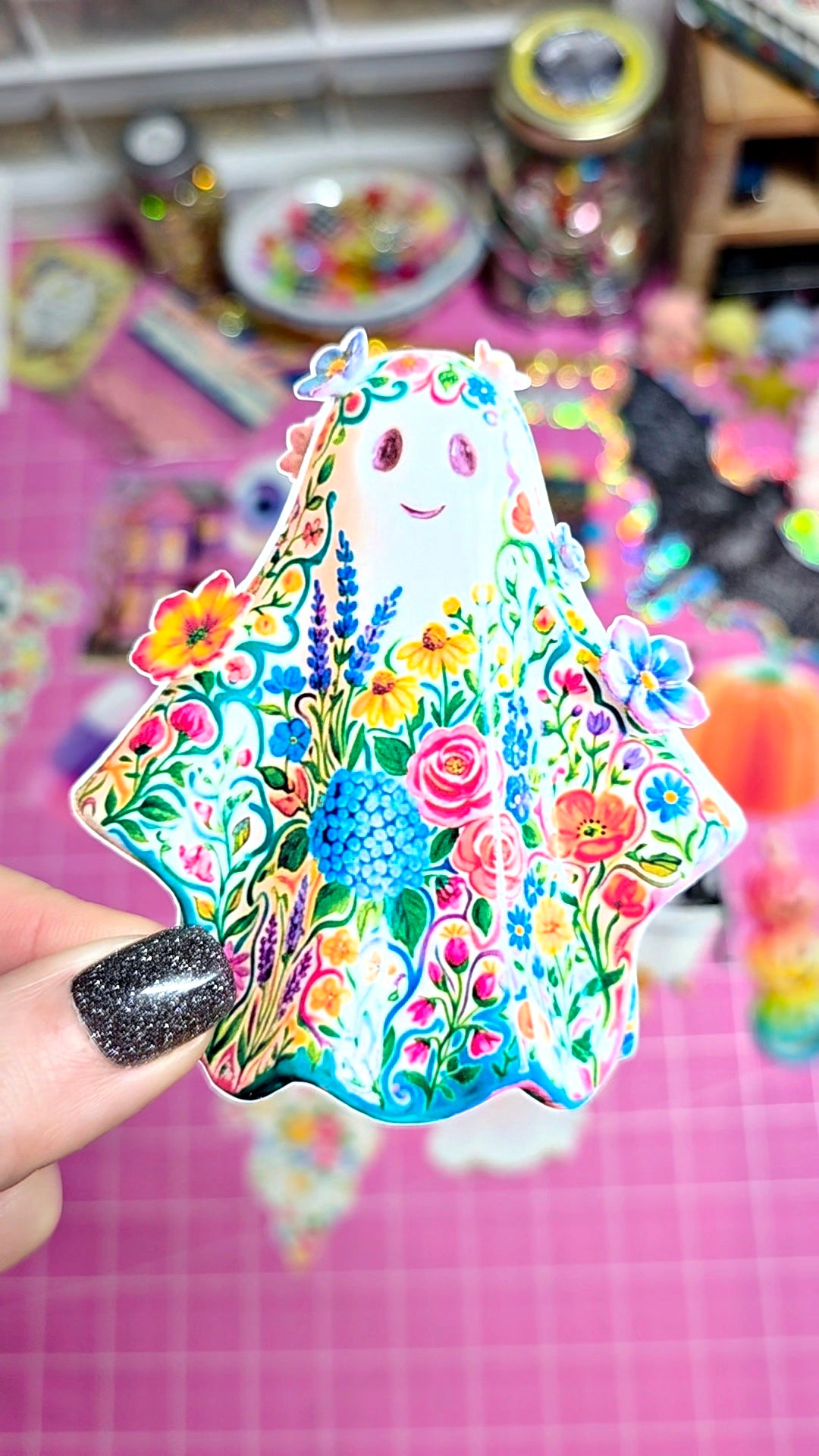 A hand with glittery black nail polish holds a colorful, ghost-shaped sticker or decal decorated with a dense floral pattern. The background is a pink cutting mat with other craft supplies blurred in the distance.