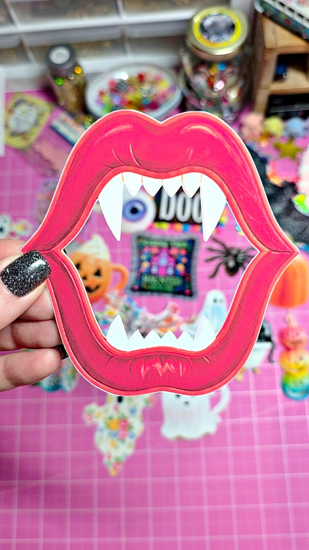 A hand with black glitter nail polish holds a pink lip-shaped mirror with white fangs, reflecting a colorful Halloween-themed background.