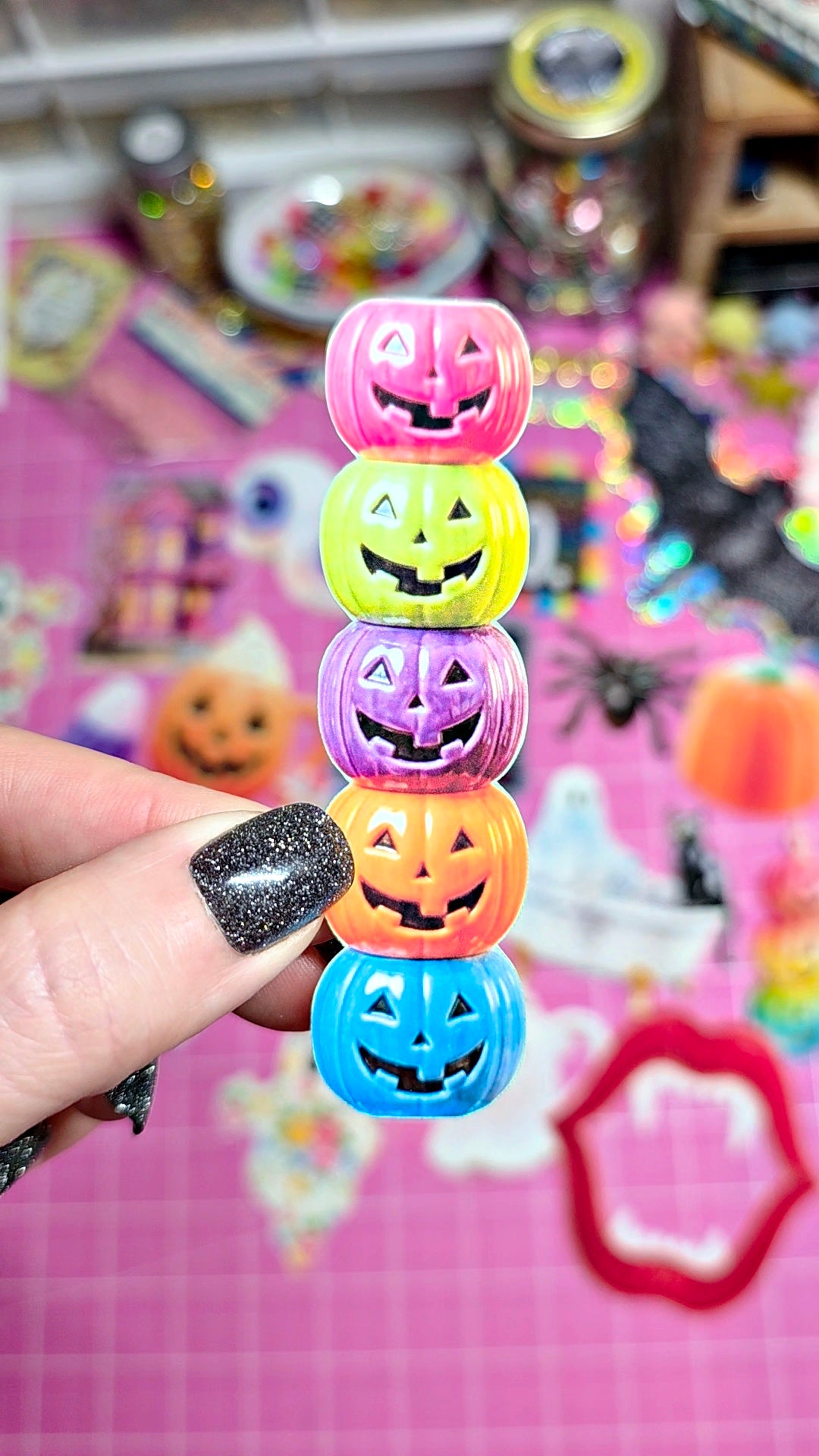 A hand with glittery black nail polish holds a stack of five colorful jack-o'-lantern-shaped rings or beads in pink, yellow, purple, orange, and blue, set against a pink craft mat background filled with Halloween decorations.