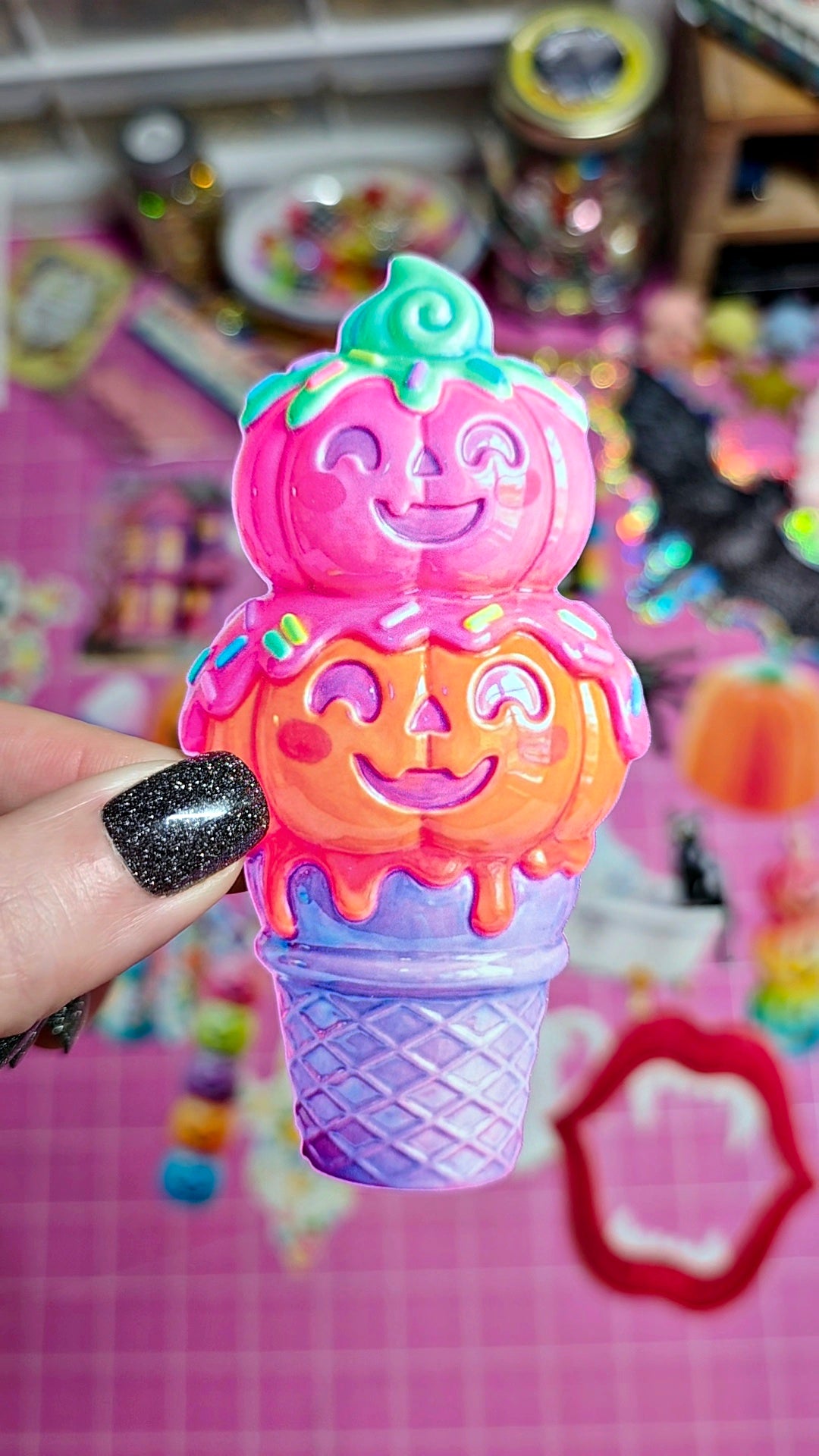 A hand with black glitter nail polish holds a colorful, jack-o'-lantern-themed ice cream toy. The toy features three stacked scoops of ice cream in pink, orange, and purple, topped with a smiling green pumpkin and dripping candy details, set against a busy, pink-themed background.