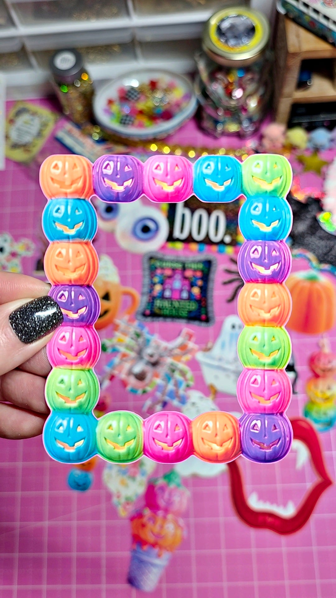 A hand with black glitter nail polish holds a rectangular plastic frame made of colorful, pumpkin-face decorated beads. The background is a busy, pink-themed craft or Halloween display with various decorations, jars, and stickers.