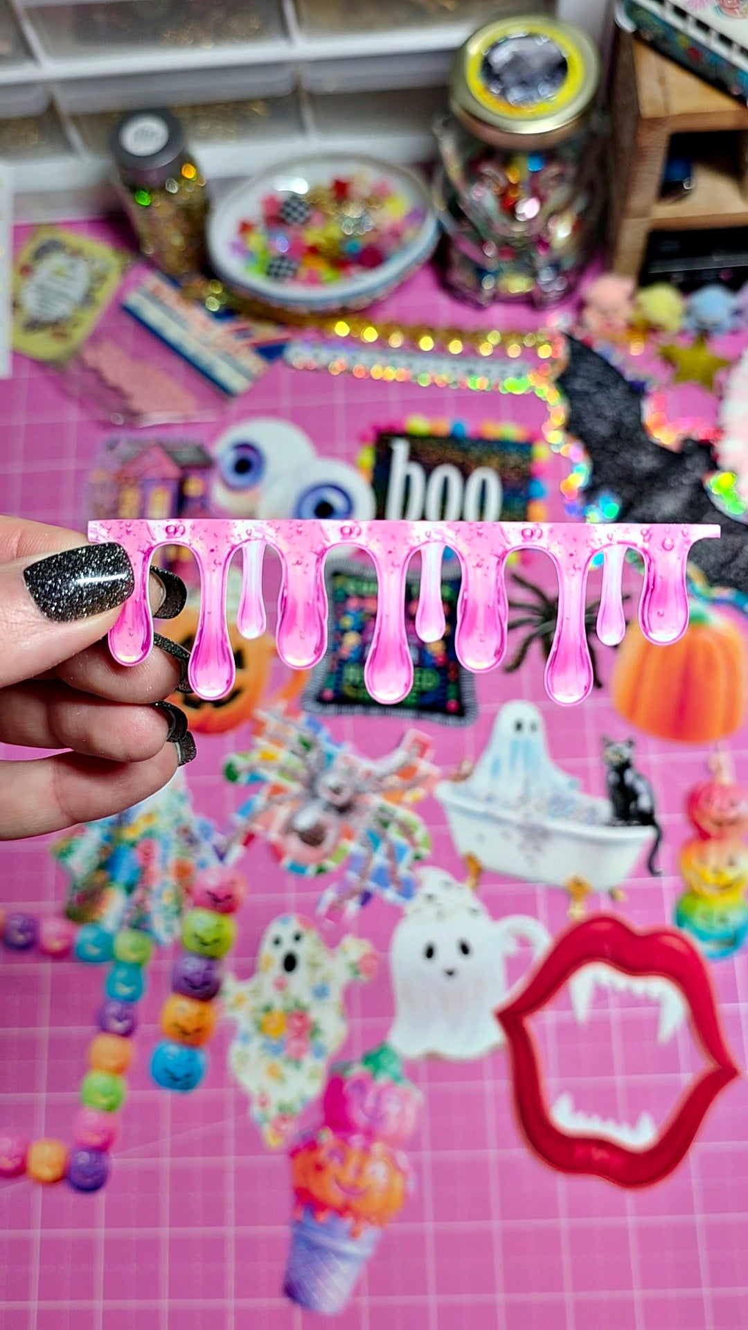 A hand with black glitter nail polish holds a pink drip-shaped sticker or decal against a busy pink craft background filled with Halloween-themed decorations, beads, and stickers.