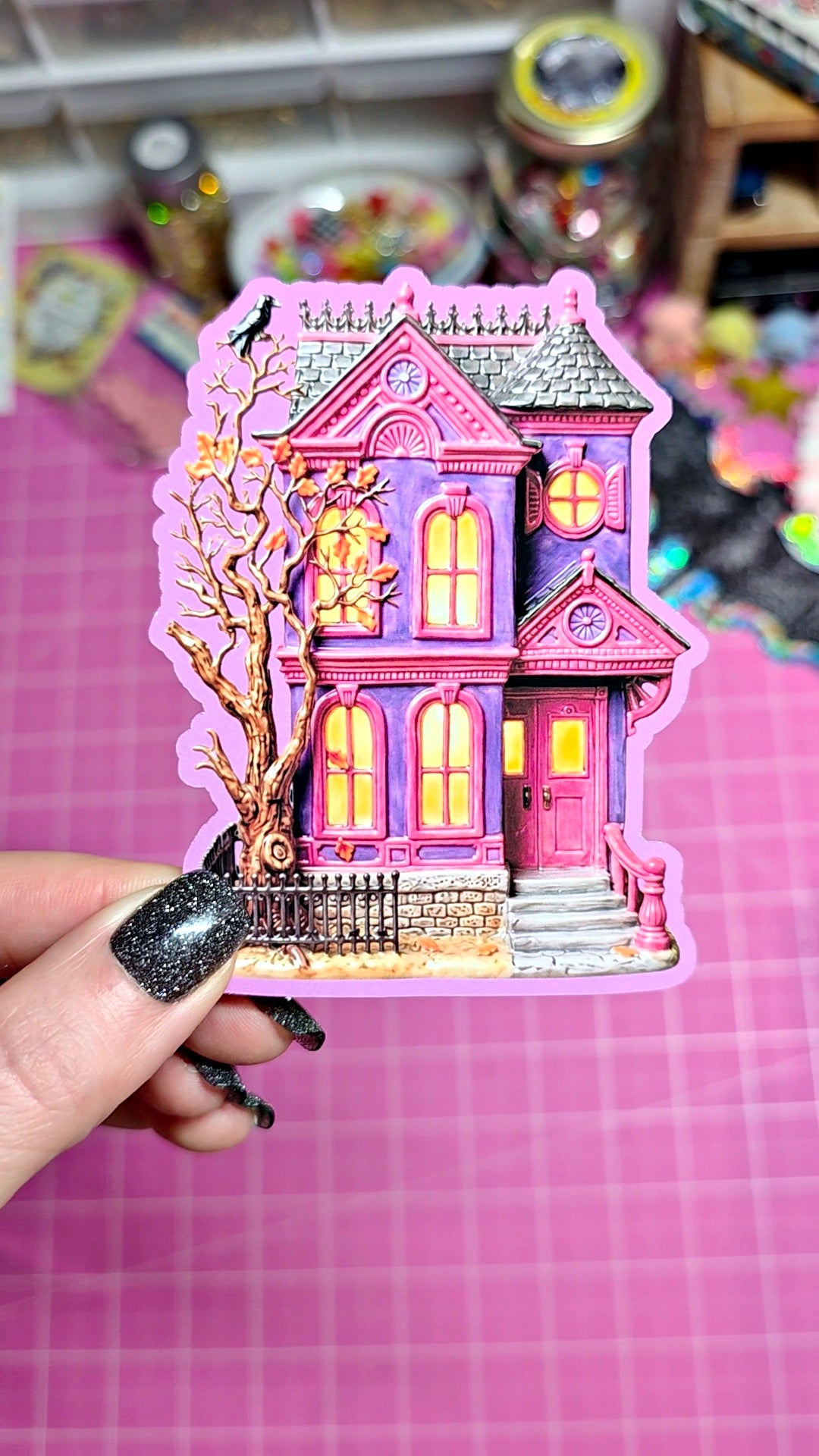 A hand with black glitter nail polish holds a die-cut sticker of a whimsical pink and purple haunted house with glowing yellow windows, set against a pink cutting mat background.