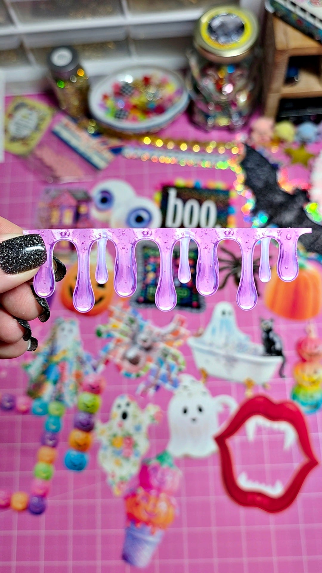 A hand with black glitter nail polish holds a pink strip of purple dripping slime above a pink craft mat covered in Halloween-themed stickers and decorations.