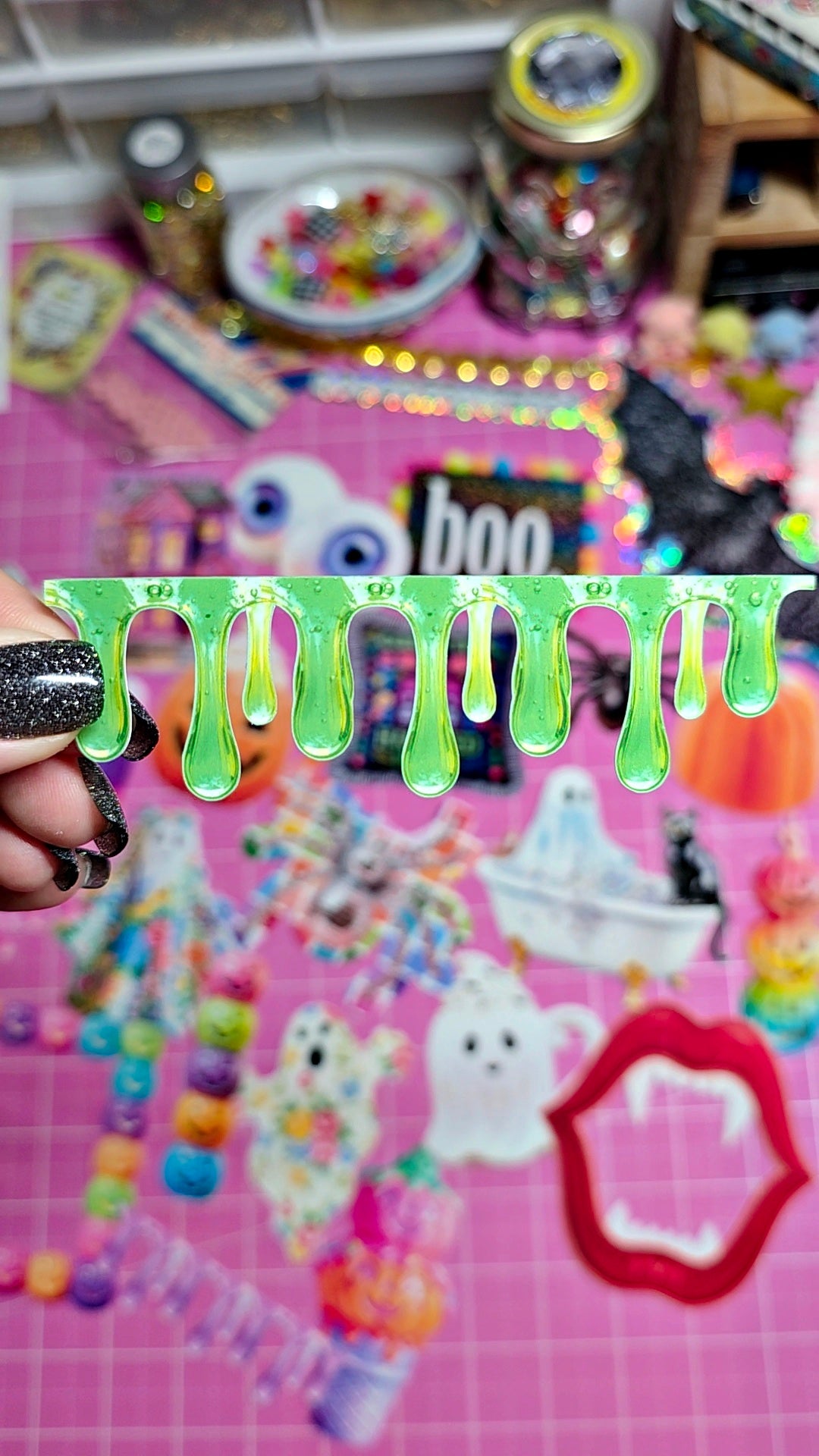 A hand with black glitter nail polish holds a green slime drip sticker against a pink craft mat covered in Halloween-themed stickers and supplies.