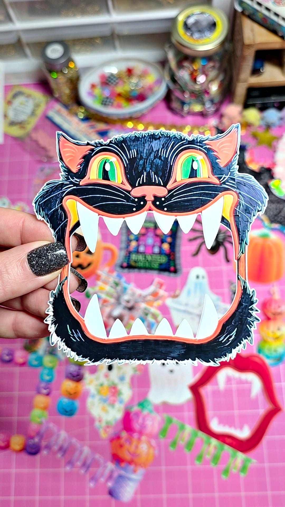 A hand with glittery black nail polish holds a die-cut black cat face sticker or craft cutout with green eyes, orange ears, and a wide open mouth showing white teeth. The background is a pink craft mat with colorful beads, jars, and Halloween-themed decorations.