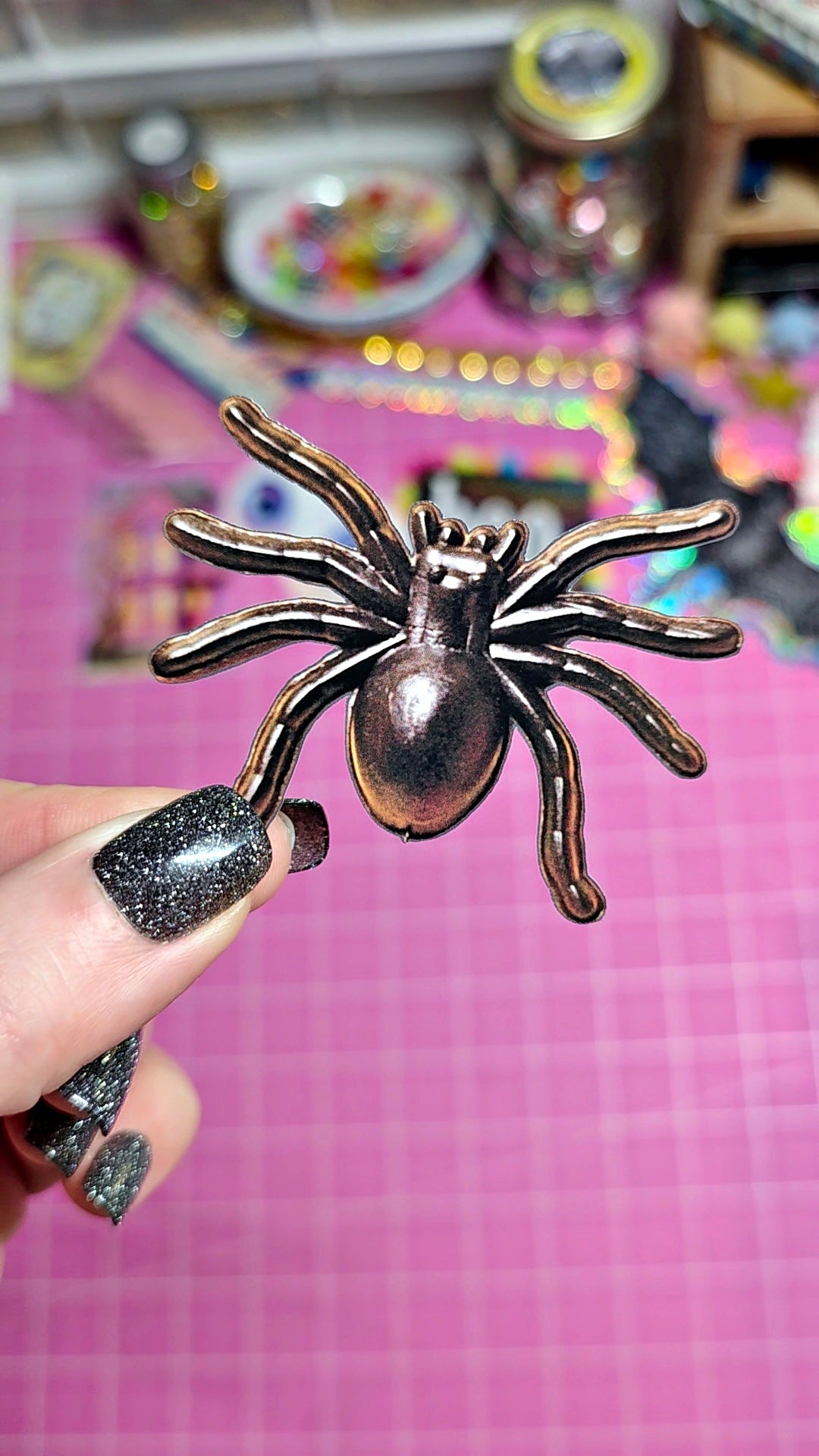 A hand with glittery black nail polish holds a metallic bronze spider-shaped hair claw clip against a pink grid cutting mat background.