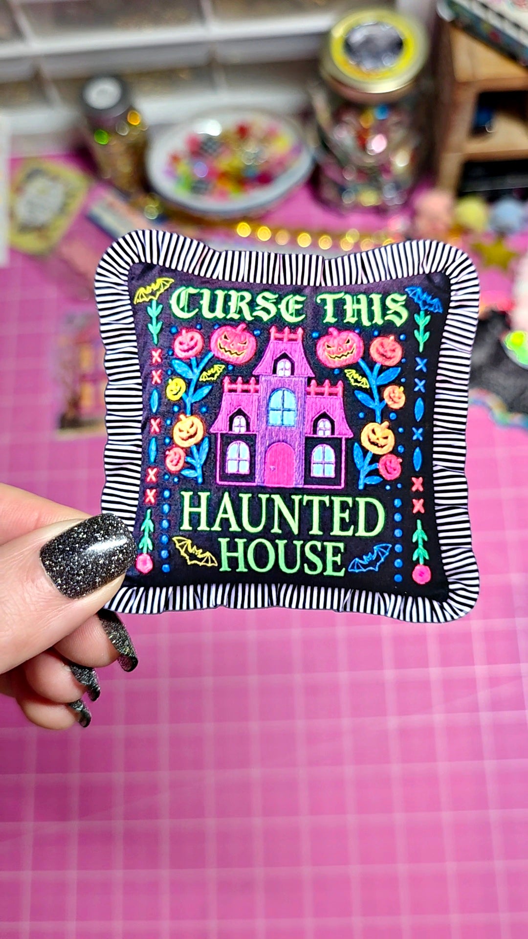 A hand with glittery black nails holds a square embroidered patch featuring a haunted house design with the text 'CURSE THIS HAUNTED HOUSE' in green and pink lettering, set against a pink cutting mat background.