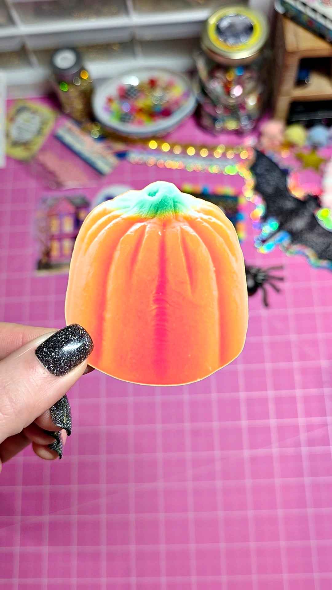 A hand with glittery black fingernails holds a small, orange pumpkin-shaped squishy toy with a mint green stem. The background is a pink grid cutting mat with various colorful Halloween decorations, jars, and accessories blurred in the background.