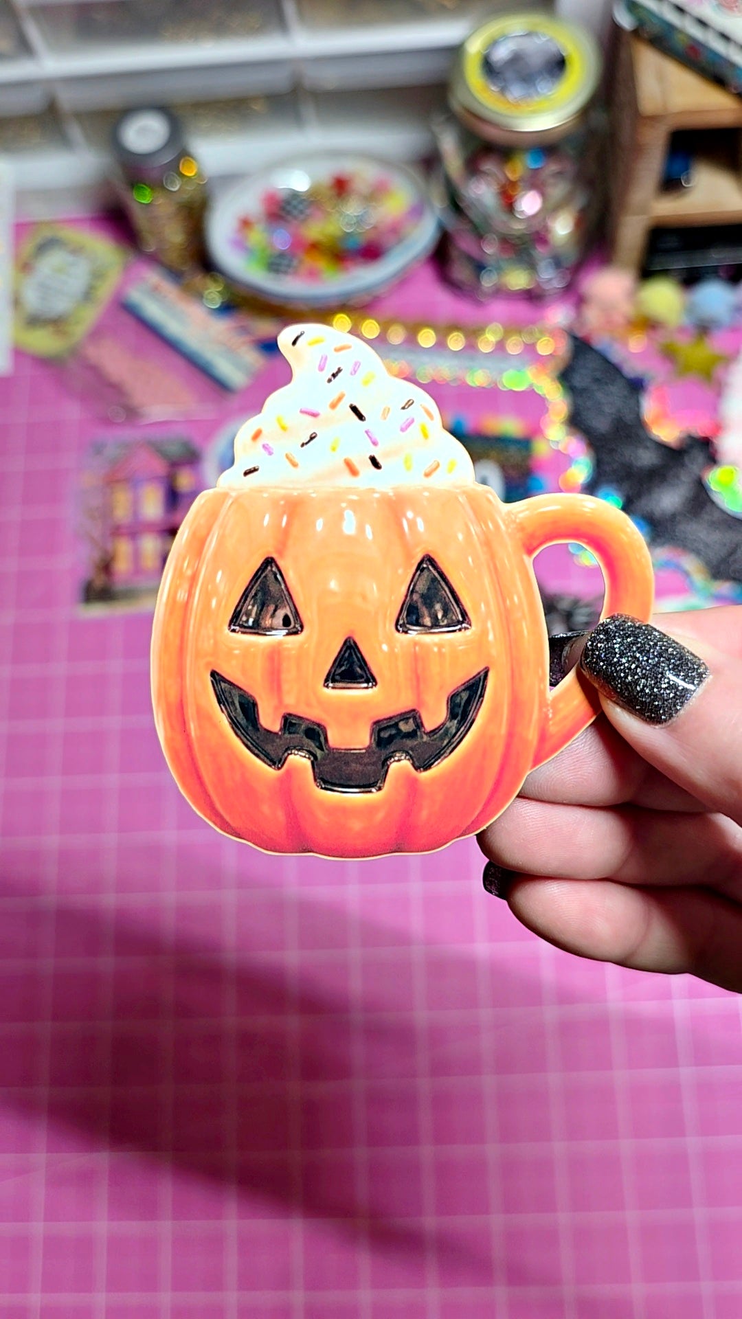 A hand with glittery black nail polish holds a small orange jack-o'-lantern mug filled with white whipped cream, set against a pink grid cutting mat and a colorful craft background.
