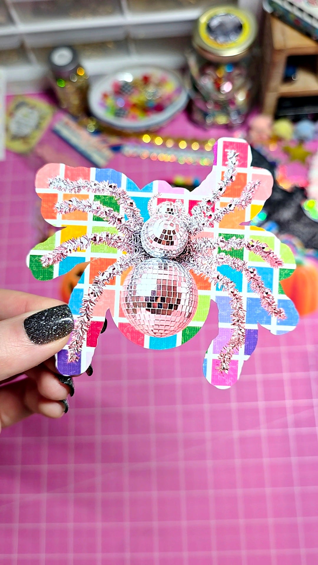 A hand with glittery black nails holds a colorful butterfly-shaped hair clip decorated with a disco ball motif, set against a pink cutting mat background with craft supplies.