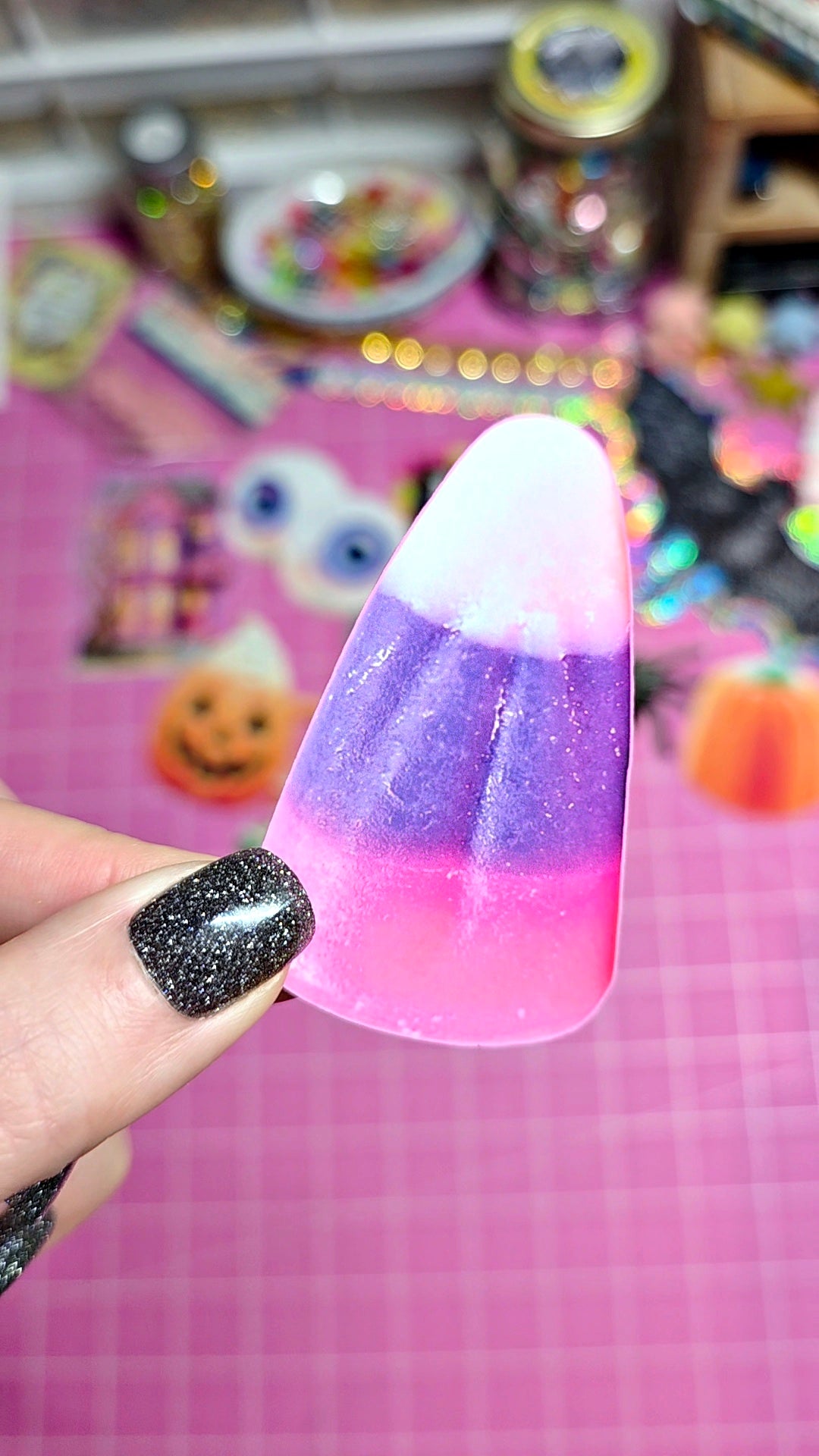 A hand with glittery black fingernails holds a candy corn-shaped nail art sticker or decal with a pink, purple, and white gradient design against a pink craft background.
