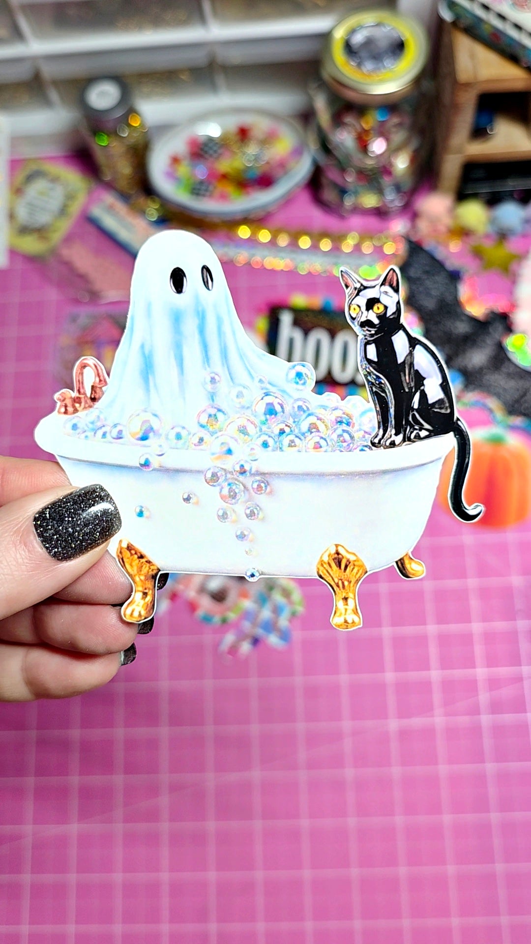 A hand with black glitter nail polish holds a small Halloween-themed enamel pin or accessory featuring a white ghost in a clawfoot bathtub with a black-and-white cat sitting on the edge, set against a pink grid background with blurred craft supplies.