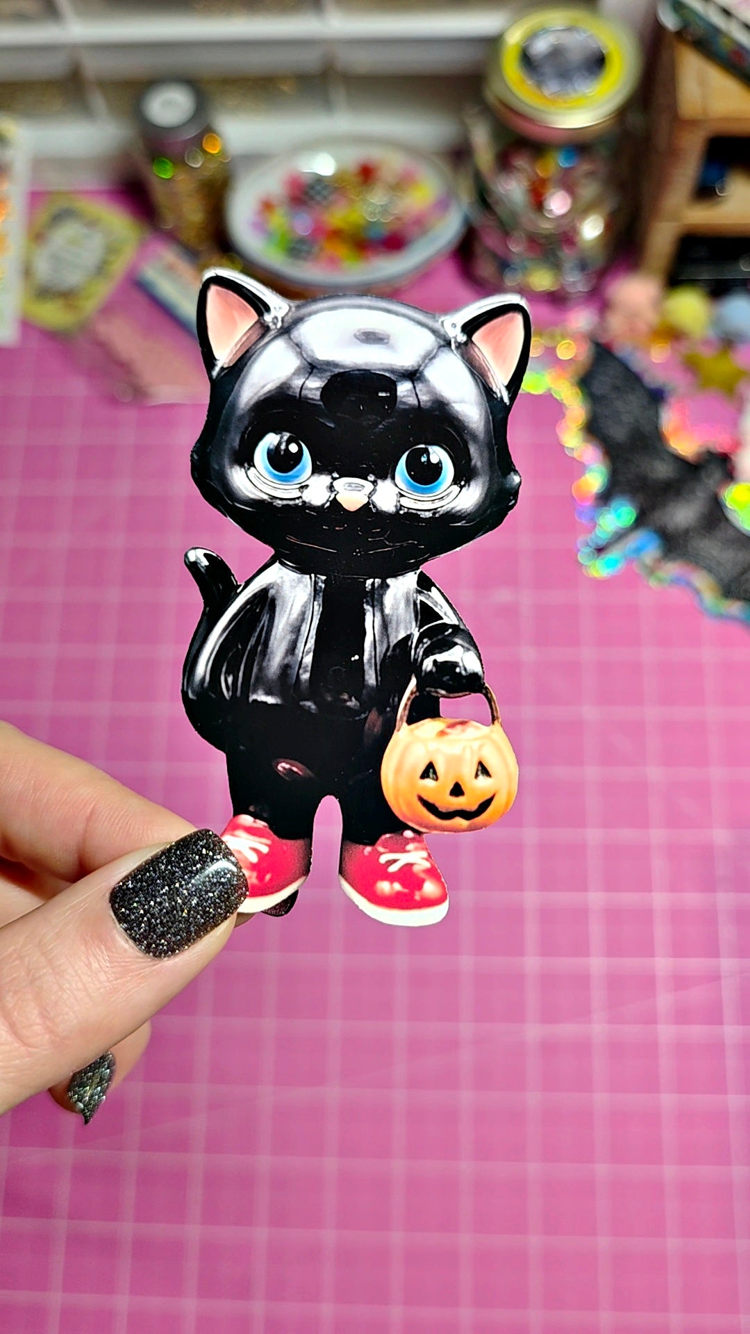 A hand with glittery black nail polish holds a small glossy black cat figurine dressed in a Halloween costume, holding a tiny orange jack-o'-lantern. The background is a pink cutting mat with colorful craft supplies blurred in the distance.