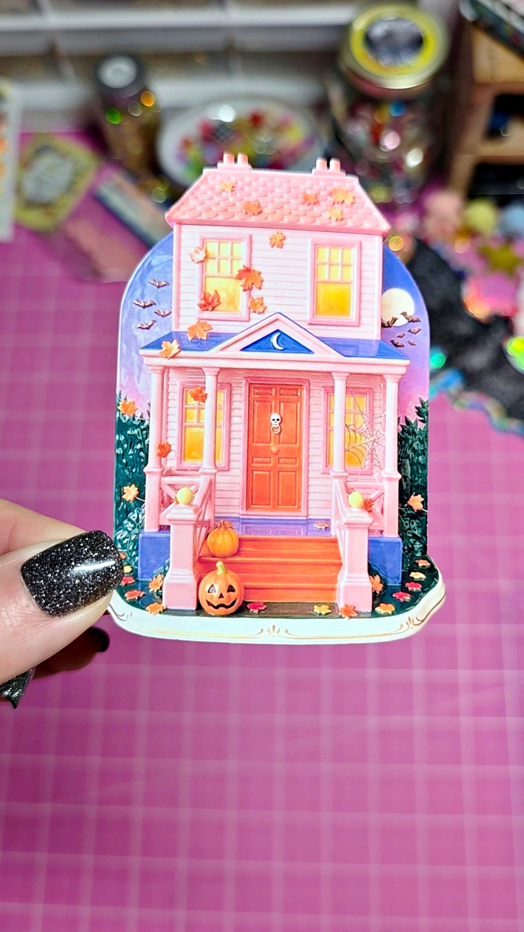 A hand with black glitter nail polish holds a small pink and orange haunted house sticker or decal against a pink grid cutting mat background.