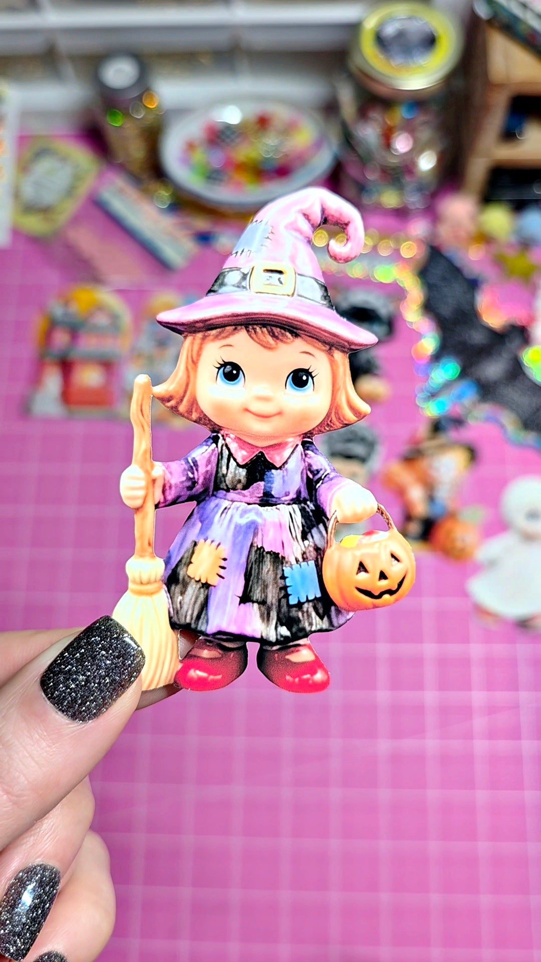 A hand with black glitter nail polish holds a small plastic doll of a witch girl in a purple and black dress with a pink hat, holding a broom and a jack-o'-lantern bucket, against a pink cutting mat background.